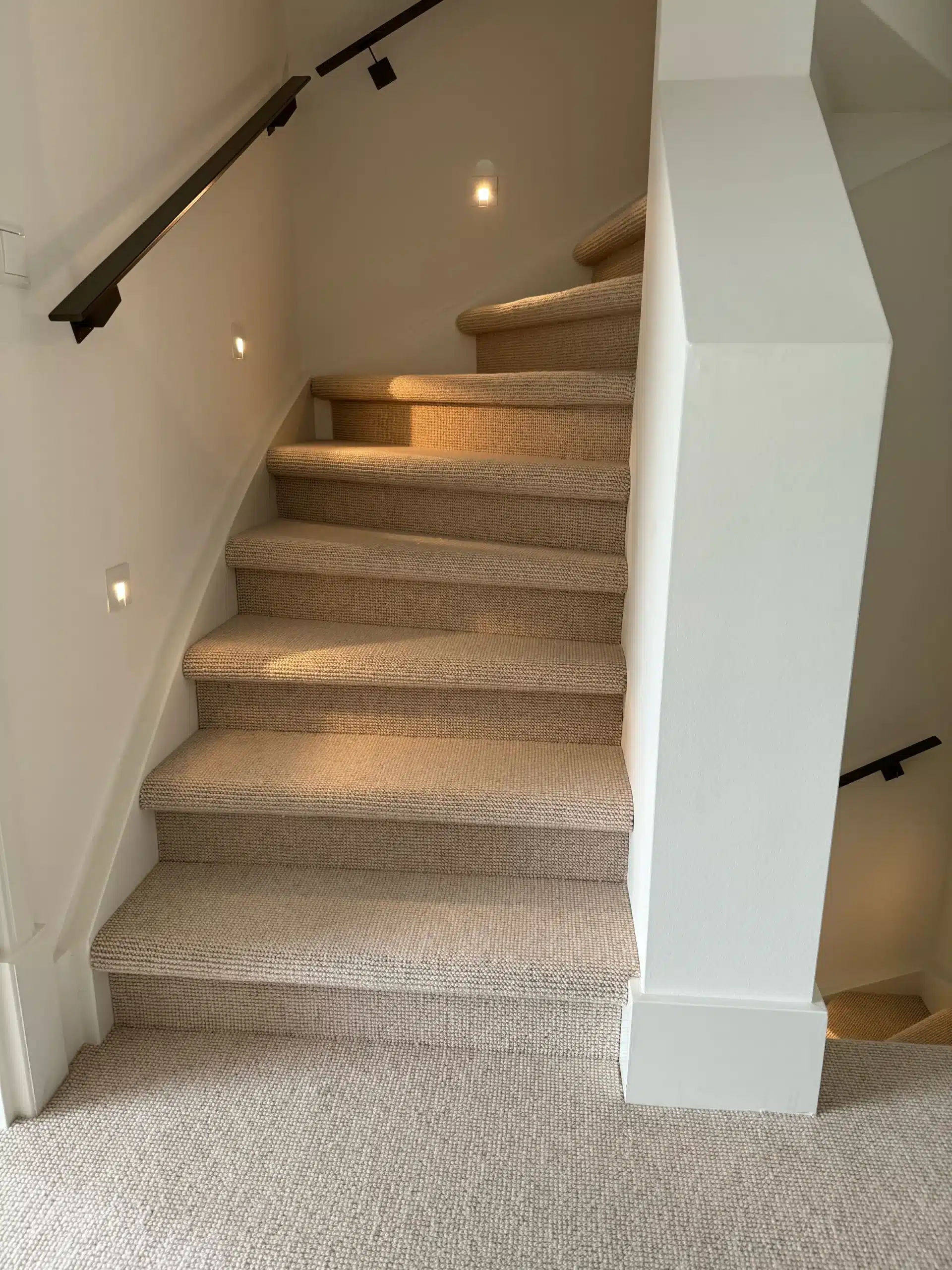 Volledig met beige wol beklede kwartslagtrap met witte wanden, donkere leuning en inbouwspots in de zijwand