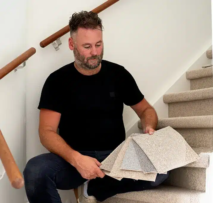 Stoffeerder bekijkt een waaier tapijtstalen in grijs en beige, zittend op een beklede trap