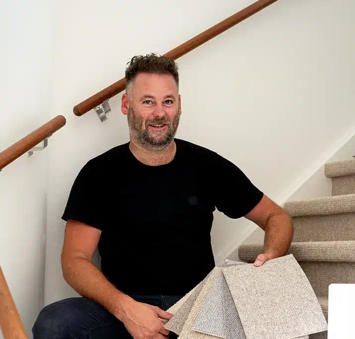 De stoffeerder achter uw wollen trap Stoffeerder in zwart shirt zit lachend op een beklede trap met een waaier tapijtstalen